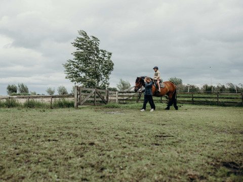 Een volwassene begeleidt een kind op een bruin paard in een weiland bij Feather Down TaarTenTuin vakantiepark.