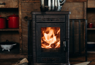 Wood stove with fire burning inside and kettle on top, logs nearby, at Feather Down TaarTenTuin kitchen.