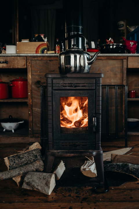 Houthaard met brandend vuur en theeketel erbovenop, houtblokken, bij Feather Down TaarTenTuin keuken.