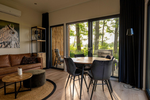 Modern lodge interior with leather sofa, dining table, large windows, and view of trees and deck outside.