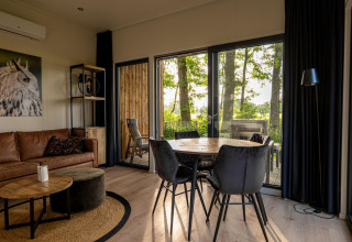 Modern lodge interior with leather sofa, dining table, large windows, and view of trees and deck outside.