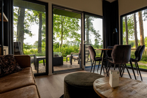 Sala de estar de una cabaña con puertas de cristal al bosque, terraza y comedor moderno con luz natural.