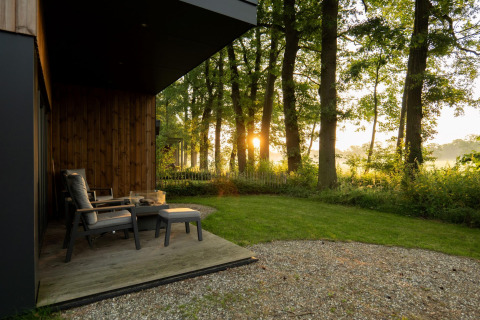 Solopgang bag træer set fra en hyggelig overdækket terrasse ved en lodge med komfortable stole.