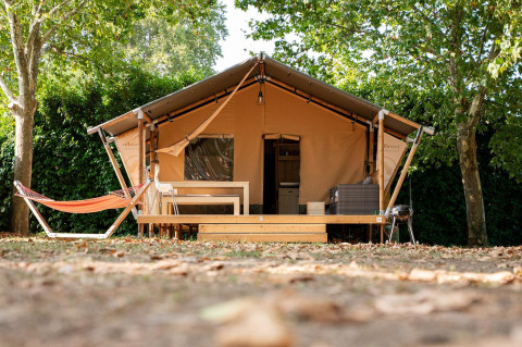 Tienda safari Villatent Nomad con terraza de madera y hamaca en Camping La Garenne en Francia, rodeada de árboles.