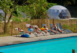 Børn slapper af på liggestole ved en pool med geodætiske kuppeltelte i baggrunden på en feriepark.