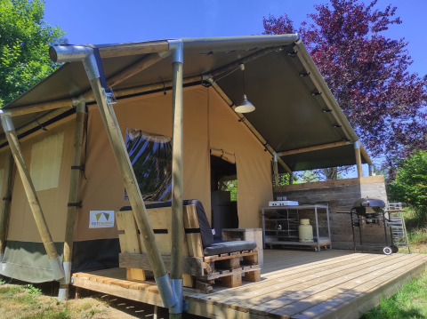Safari tent with a wooden deck, outdoor kitchen, and grill set up, surrounded by greenery and sunlight.