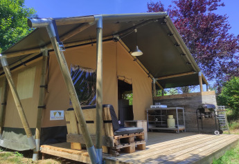 Safari tent with a wooden deck, outdoor kitchen, and grill set up, surrounded by greenery and sunlight.