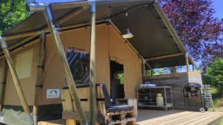 Et billede af et safaritelt med træterrasse, udekøkken og grill, omgivet af skov og solskin.