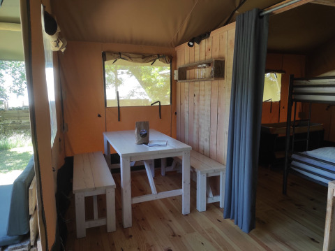 Vue intérieure d'une tente safari avec table en bois, bancs, lit superposé et murs en bois éclairés.