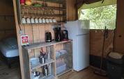 Pequeña cocina de tienda safari con estantes de madera, nevera, cafetera y vajilla a la vista.
