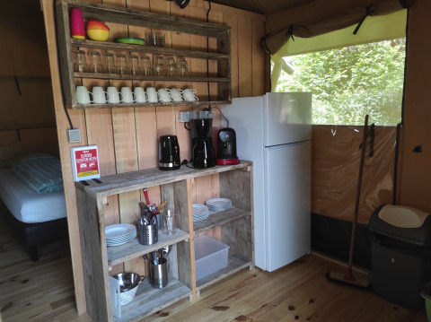 Pequeña cocina de tienda safari con estantes de madera, nevera, cafetera y vajilla a la vista.
