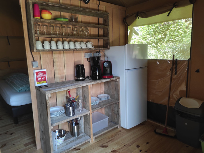 Pequeña cocina de tienda safari con estantes de madera, nevera, cafetera y vajilla a la vista.