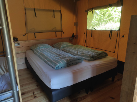 Vista interna di una tenda safari con letto matrimoniale, lenzuola a righe e finestre sulla natura.