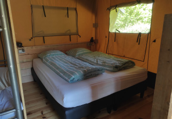 Vista interna di una tenda safari con letto matrimoniale, lenzuola a righe e finestre sulla natura.