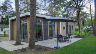 Moderne lodge Cube la Mer in De Zanding, Nederland, omringd door bomen en een terras met tuinmeubilair.