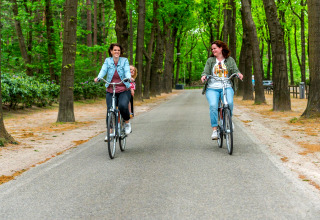 Two women ride bicycles on a forest path at Cube la Mer, De Zanding, Netherlands, with one carrying a child.