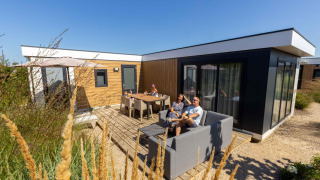 Famiglia che si rilassa all'esterno di Zonneweelde Lodge XL al Camping Zonneweelde nei Paesi Bassi in una giornata soleggiata.
