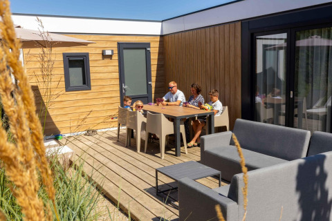 Famille se détendant sur la terrasse de la Zonneweelde Lodge XL au Camping Zonneweelde, aux Pays-Bas, par beau temps.