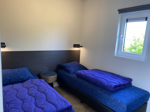 Chambre avec deux lits simples, literie bleue, table de chevet et fenêtre à Zonneweelde Lodge XL, Camping Zonneweelde.