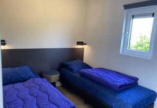 Twee éénpersoonsbedden, blauwe dekbedden, nachtkastje en raam in Zonneweelde Lodge XL op Camping Zonneweelde.