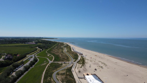Vue aérienne d'une plage près d'un lodge avec cabanes, sentiers et espaces verts sous un ciel bleu clair.
