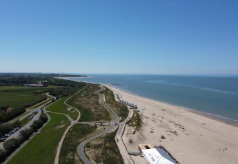 Veduta aerea di una spiaggia presso un lodge con cabine, sentieri e prati verdi sotto un cielo blu chiaro.