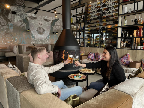 Een stel geniet van eten en drinken bij de open haard in een moderne lodge met muurschildering en wijnrek.