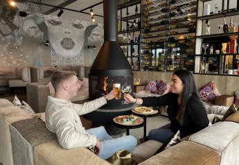 Una coppia gusta cibo e bevande davanti al camino in un moderno lodge con arte murale e scaffali di vini.