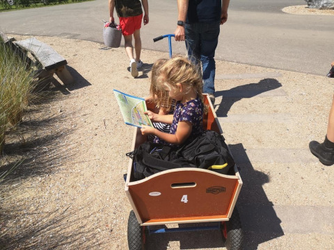 Un niño pequeño se sienta en un carro de madera leyendo un mapa mientras un adulto tira de él en Camping Zonneweelde.