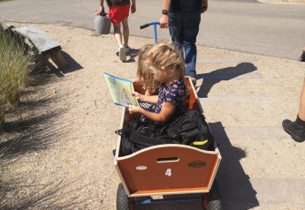 Un niño pequeño se sienta en un carro de madera leyendo un mapa mientras un adulto tira de él en Camping Zonneweelde.