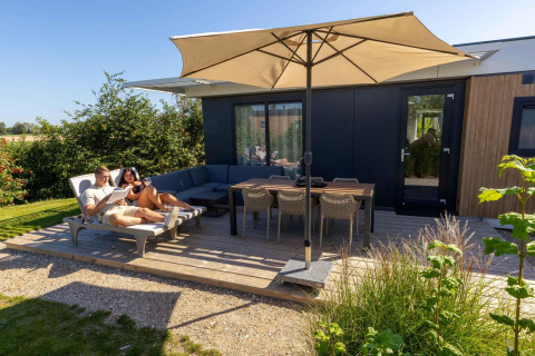 Dos personas relajándose en la terraza de Zonneweelde Lodge en Camping Zonneweelde, Países Bajos, bajo una sombrilla.