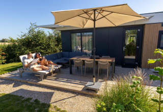 Dos personas relajándose en la terraza de Zonneweelde Lodge en Camping Zonneweelde, Países Bajos, bajo una sombrilla.