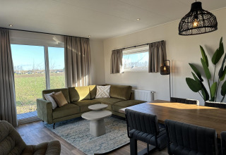 Moderne stue i Zonneweelde Lodge, Camping Zonneweelde, Holland med stort vindue, grøn sofa og spisebord.