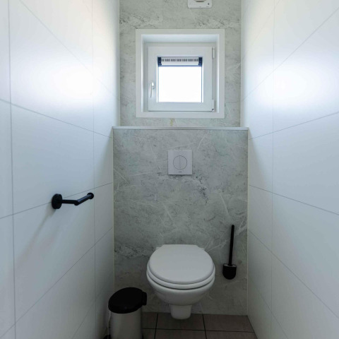 Et moderne toilet med væghængt wc, lille vindue, toiletbørste og skraldespand på Zonneweelde Lodge.