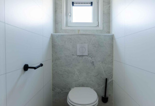 Et moderne toilet med væghængt wc, lille vindue, toiletbørste og skraldespand på Zonneweelde Lodge.