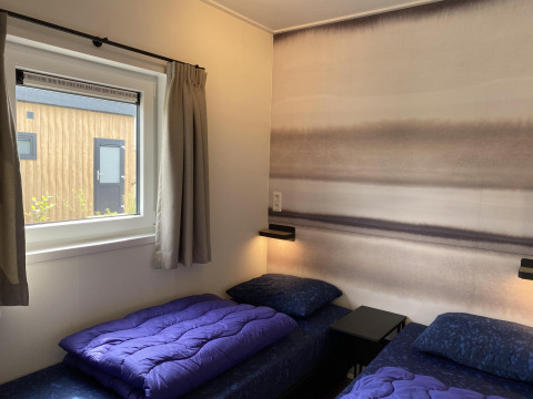 To enkeltsenge med lilla sengetøj på et moderne værelse med udsigt til træfacade, Zonneweelde Lodge.