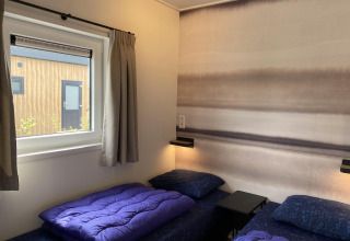 To enkeltsenge med lilla sengetøj på et moderne værelse med udsigt til træfacade, Zonneweelde Lodge.