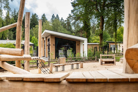 Legeplads med træstrukturer foran moderne lodges på Resort Veluwe, Silva, i Holland omgivet af skov.