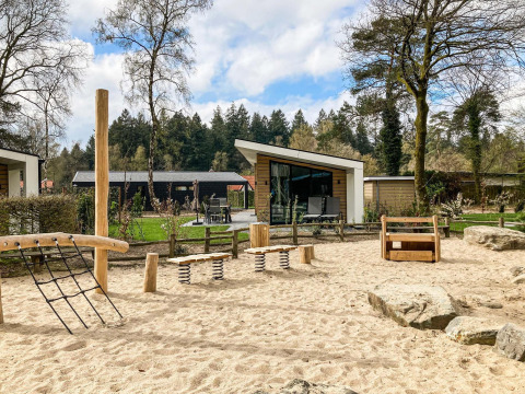 Spielplatz mit Holzausstattung und Sand vor modernen Lodges im Silva Playground, Resort Veluwe, Niederlande.