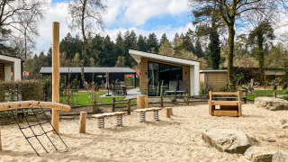 Spielplatz mit Holzausstattung und Sand vor modernen Lodges im Silva Playground, Resort Veluwe, Niederlande.
