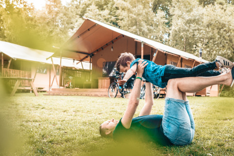 En person leger med et barn foran en Vecht Glamping Lodge safariteltscene på en græsplæne i naturen.