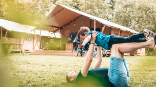 Una persona juega con un niño en el césped frente a una tienda safari Vecht Glamping Lodge rodeada de naturaleza.