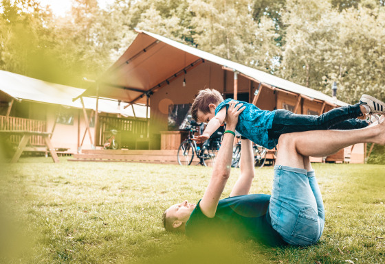 Una persona juega con un niño en el césped frente a una tienda safari Vecht Glamping Lodge rodeada de naturaleza.