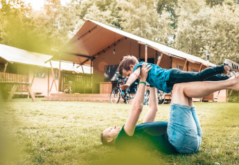 Una persona juega con un niño en el césped frente a una tienda safari Vecht Glamping Lodge rodeada de naturaleza.