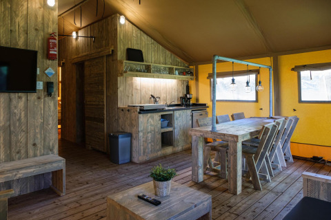 Vista interior de una tienda de glamping safari con muebles de madera, mesa de comedor y pequeña cocina.