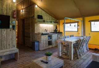 Vista interior de una tienda de glamping safari con muebles de madera, mesa de comedor y pequeña cocina.
