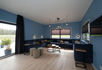Lounge van Amalia Familia-lodge in Hofparken Wiltershaar, Nederland, met blauwe muren, banken en grote ramen.