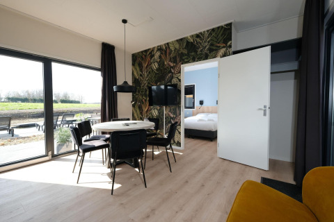 Modern lodge interior with dining table, black chairs, large windows and bedroom view at Hofparken Wiltershaar.