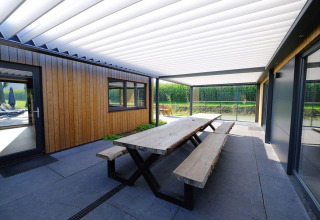 Espacio exterior con mesa y bancos de madera bajo porche en Amalia Familia, Hofparken Wiltershaar, Países Bajos.