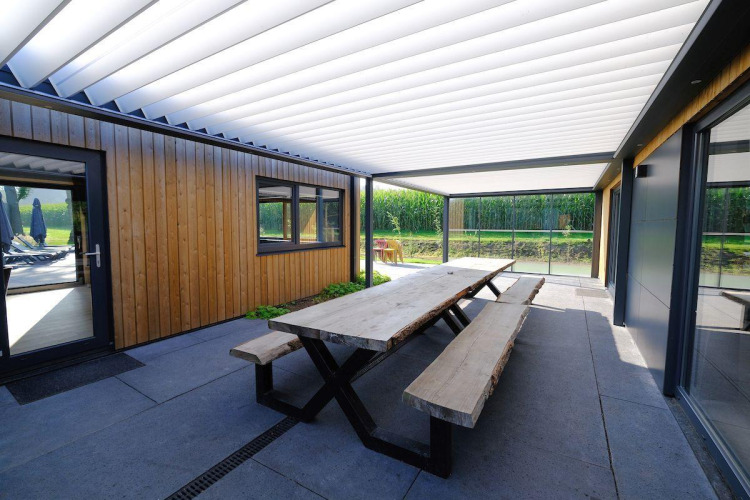 Espacio exterior con mesa y bancos de madera bajo porche en Amalia Familia, Hofparken Wiltershaar, Países Bajos.